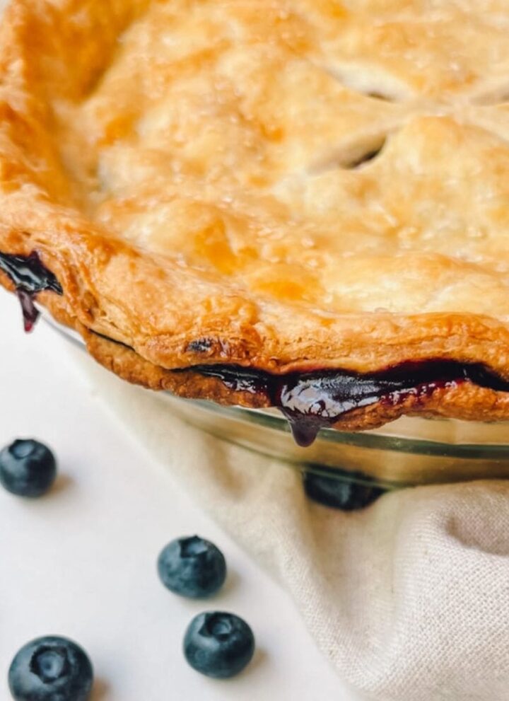 close up of blueberry pie and crust with loose blueberries surrounding pie dish