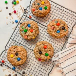 overhead picture of five cookies on a cooling rack surrounded by m&m's, chocolate chips, and oats.