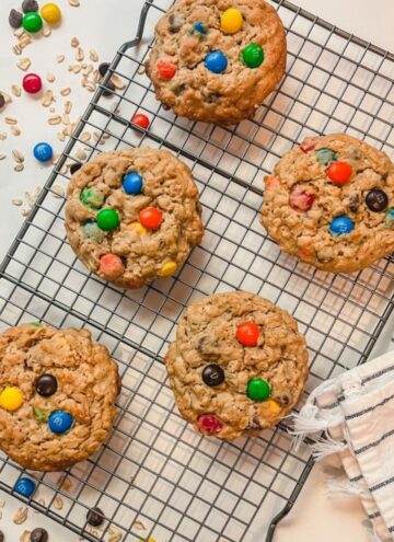 overhead picture of five cookies on a cooling rack surrounded by m&m's, chocolate chips, and oats.