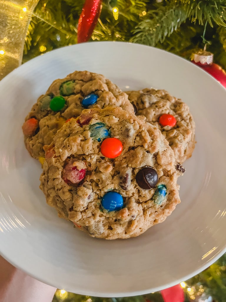 close up of three m&m monster cookies on a plate in front of a lit up Christmas tree