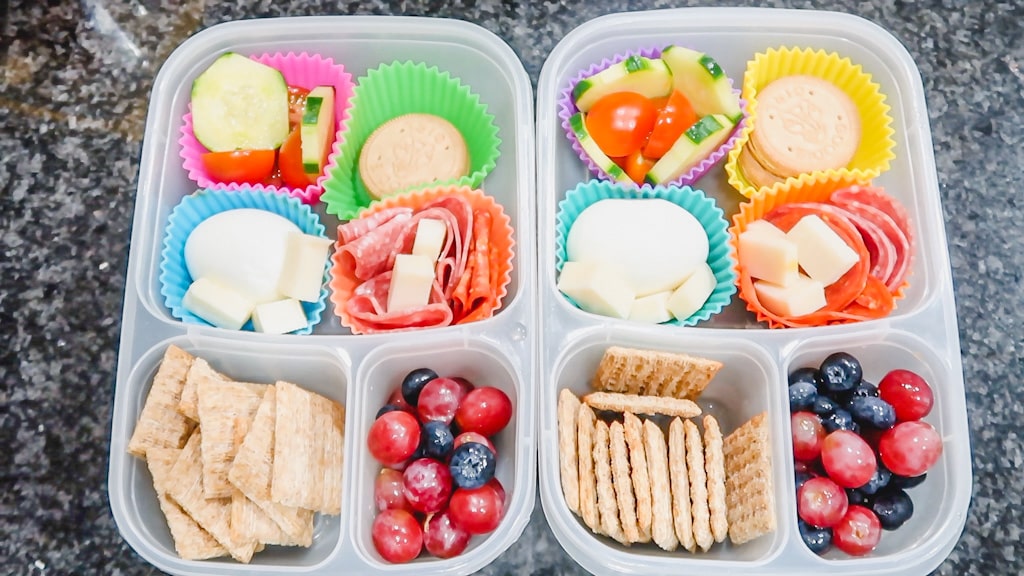 overhead picture of kids school lunch of hard boiled egg, salami, cheese, crackers, cucumbers, tomatoes, grapes, and triscuit crackers