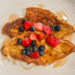 croissant french toast on a white plate with strawberries, blueberries and maple syrup on top