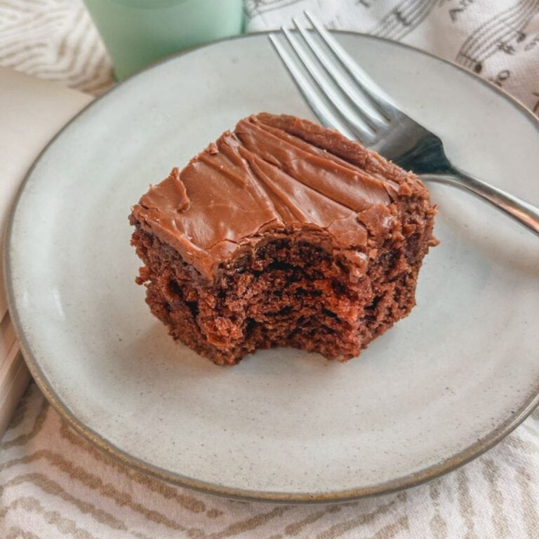 close up of a slice of brownie cake with a bite taken out of the corner on a white plate with a fork behind the cake.