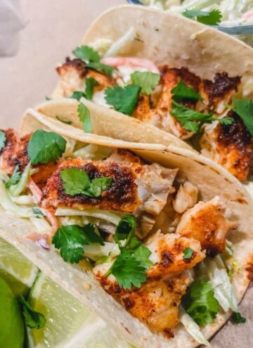 the best baked fish taco recipe with tacos on top of cutting board with lime wedges and broccoli slaw next to the tacos