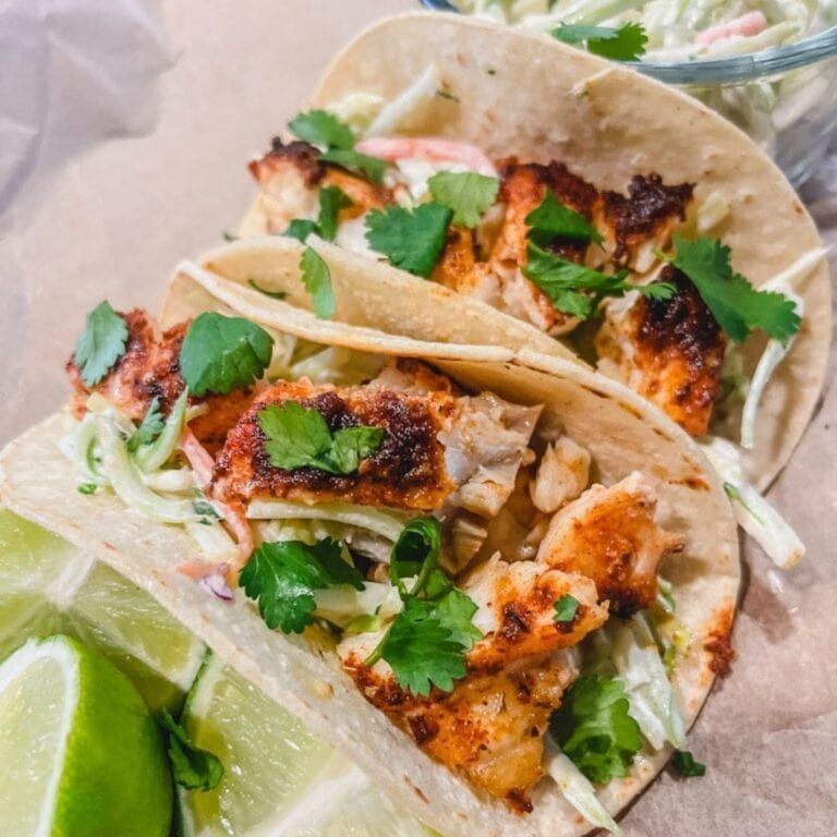 the best baked fish taco recipe with tacos on top of cutting board with lime wedges and broccoli slaw next to the tacos