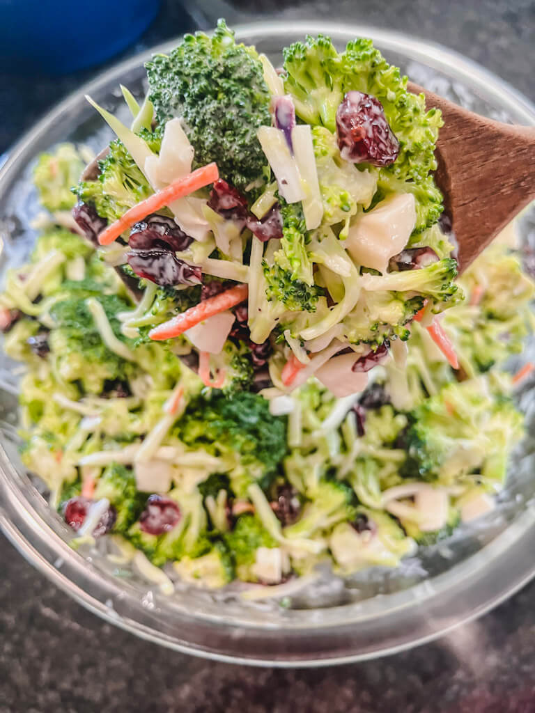 close up picture of broccoli apple salad on a wooden spoon over a clear glass bowl.