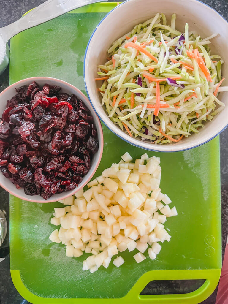 bowls of broccoli slaw, dried cranberries, and diced honeycrisp apple on a green cutting board.