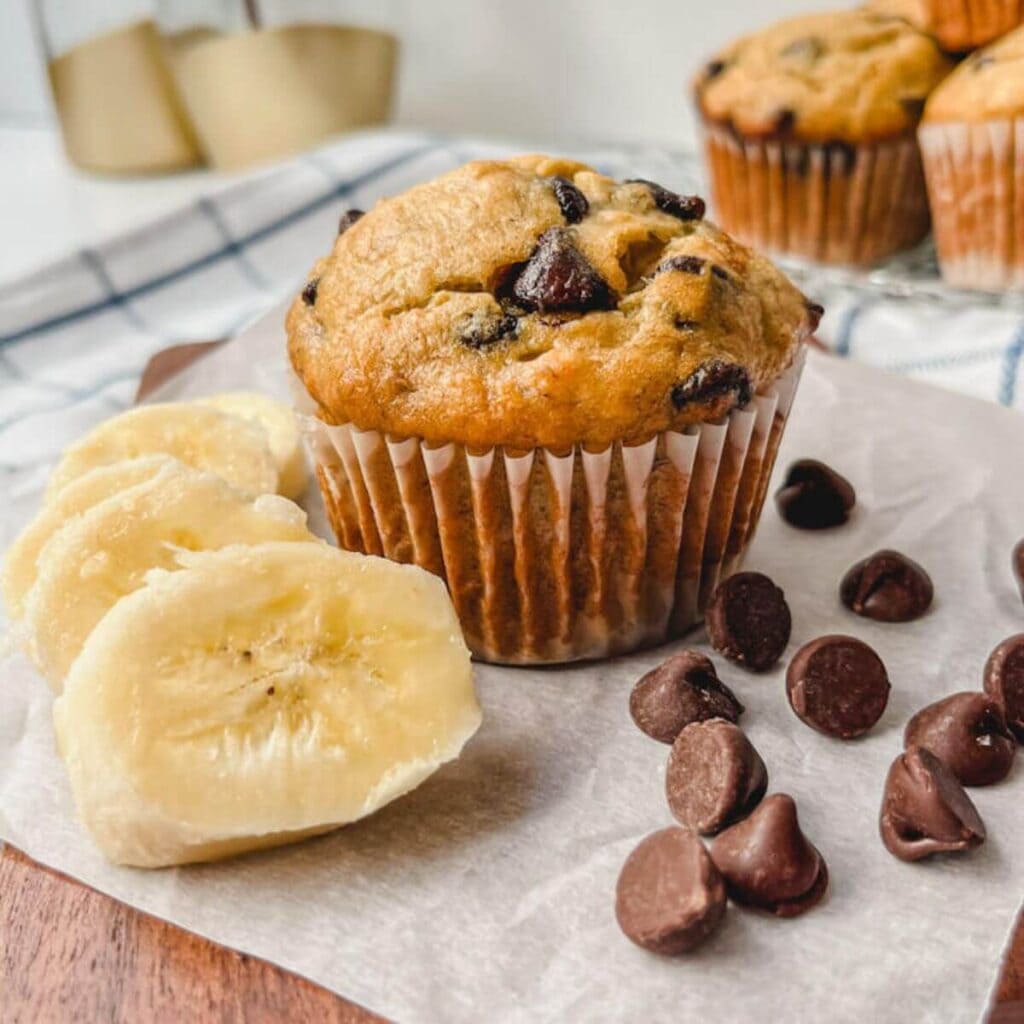 close up of dark chocolate chip banana muffin with slices of bananas and chocolate chips around the muffin on a parchment paper.