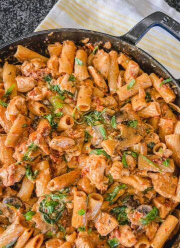 overhead image of one pot sun-dried tomato and chicken pasta in a large cast iron skillet with a a yellow and white towel next to the skillet.