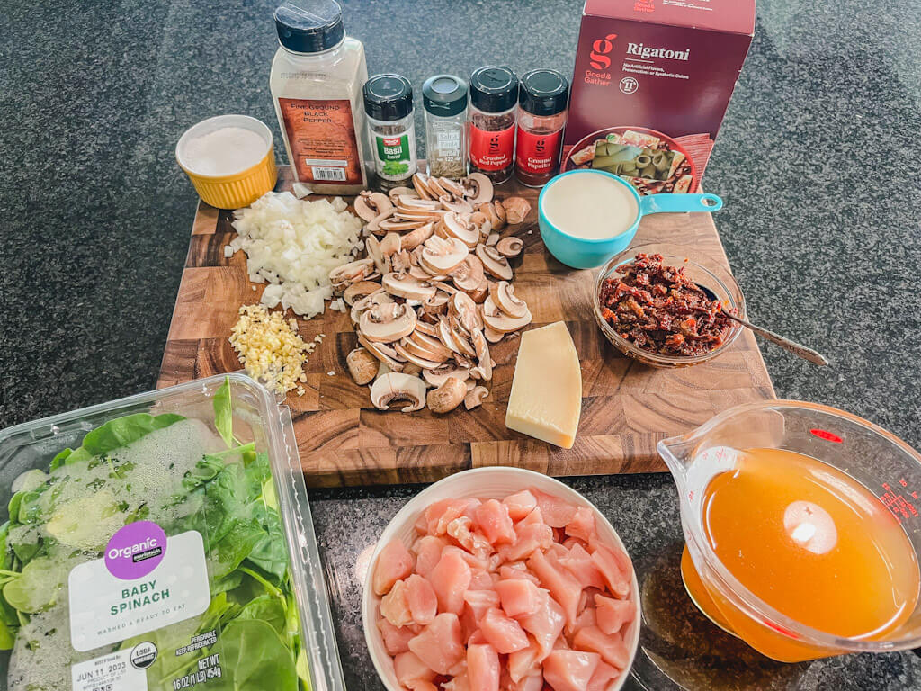 ingredients laid out on a wooden cutting board for a one-pot sun-dried tomato and chicken pasta recipe.