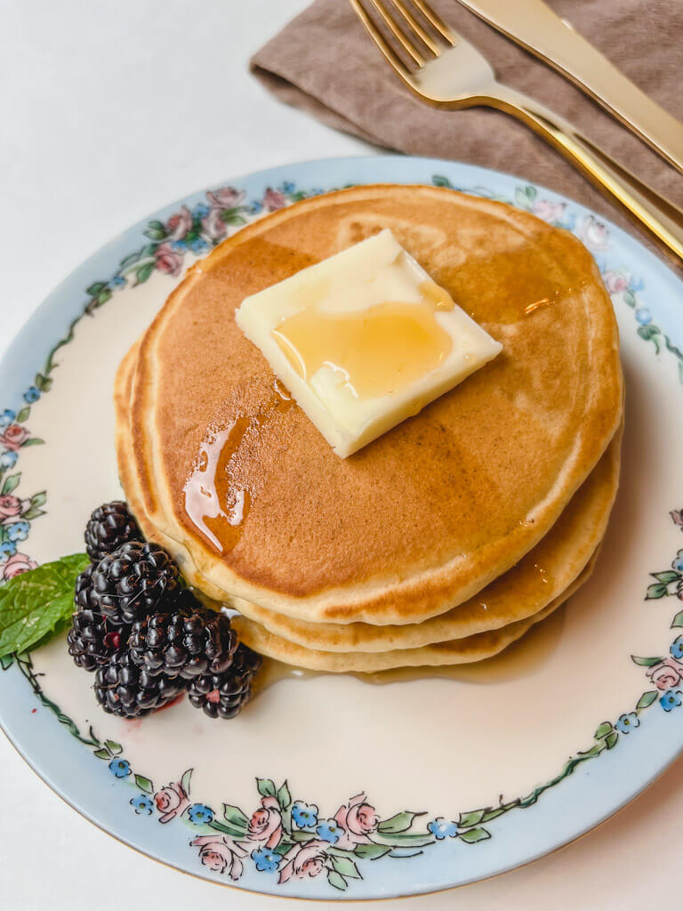 overhead picture of 3 pancakes with a slice of butter and maple syrup drizzled over pancakes on a plate with a handful of blackberries next to them.