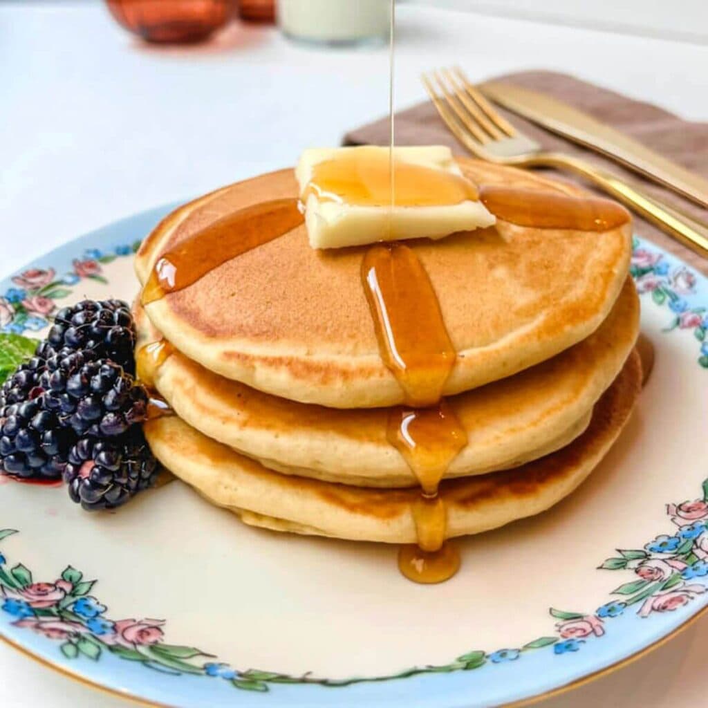 close up of 3 pancakes stacked on top of each other on a plate with butter and syrup.