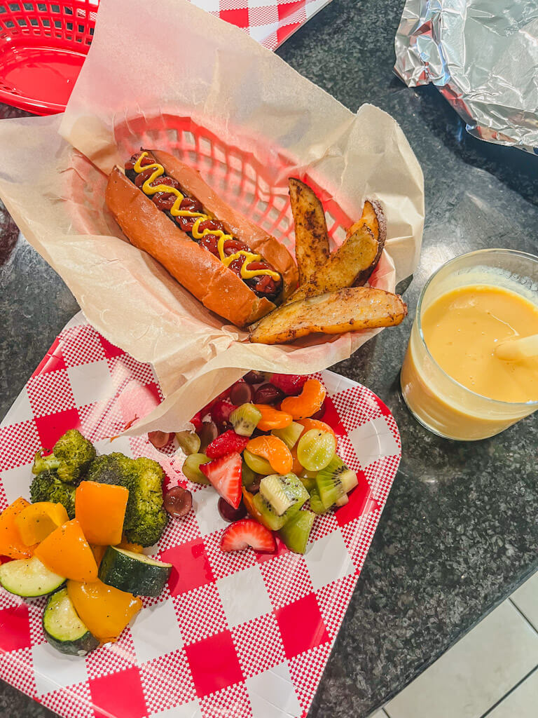 a picture of a hot dog and potato wedges in a basket with a glass of orange julius next to the basket and a plate of fruit salad and vegetables on the counter. 