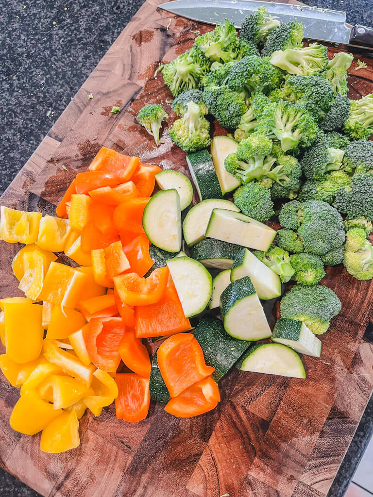 orange and yellow bell peppers, zucchini and broccoli cut up on a wooden chopping board.