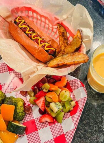 overhead image of a hot dog with potato wedges, roast vegetables, a fruit salad, and an orange julius on a black countertop for an Oliver and Company Dinner night
