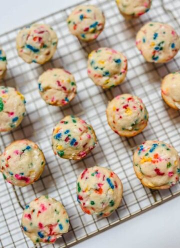 16 mini funfetti muffins on a gold wire rack on top of a white cardboard sheet.