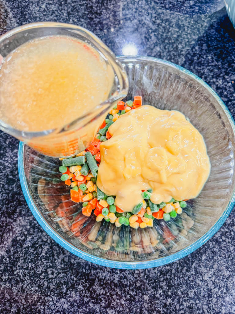 a measuring cup holding chicken broth over the bowl of frozen vegetables and cream of chicken soup. 