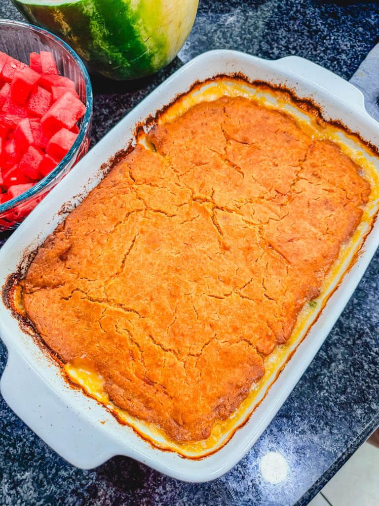 overhead view of the casserole resting in the white baking dish next to a bowl of sliced watermelon on a black counter top.
