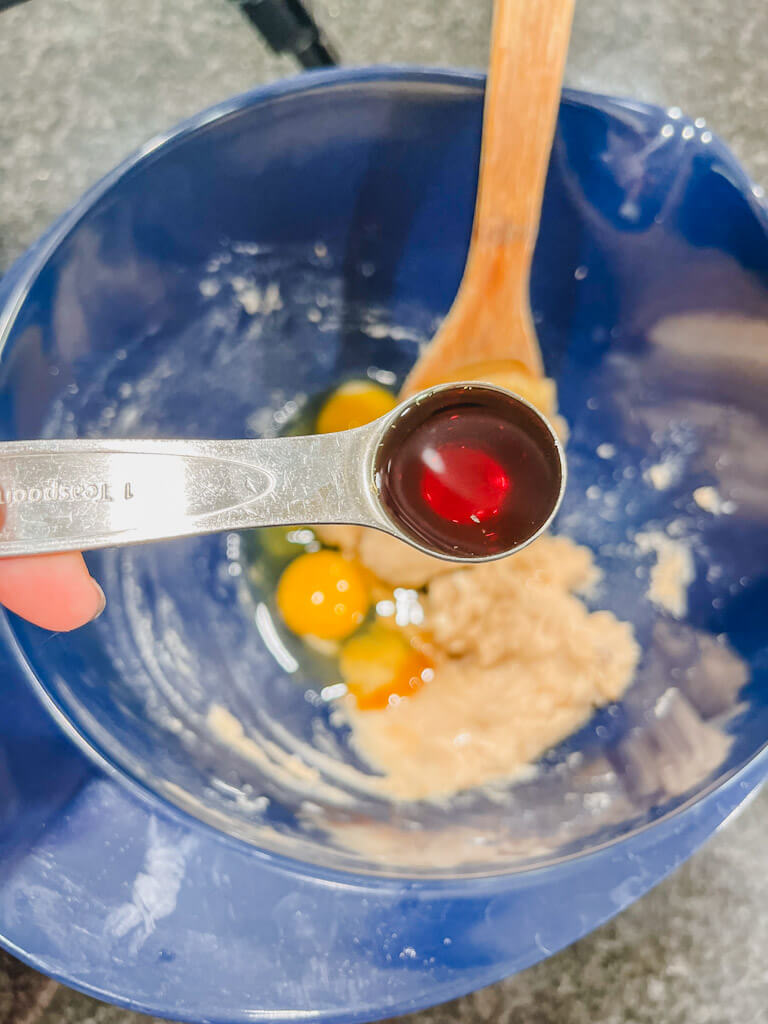 vanilla extract in a teaspoon being held over the blue mixing bowl along with two cracked eggs in the bowl.
