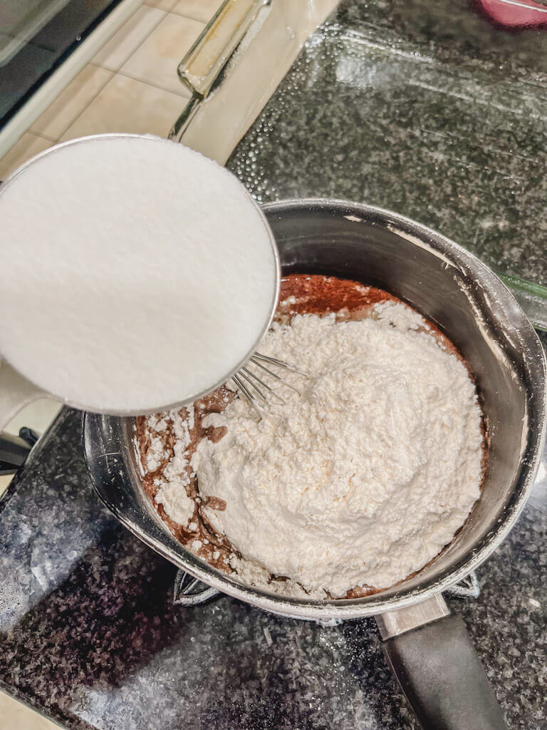 sugar in a measuring cup being added to the flour in a saucepan.