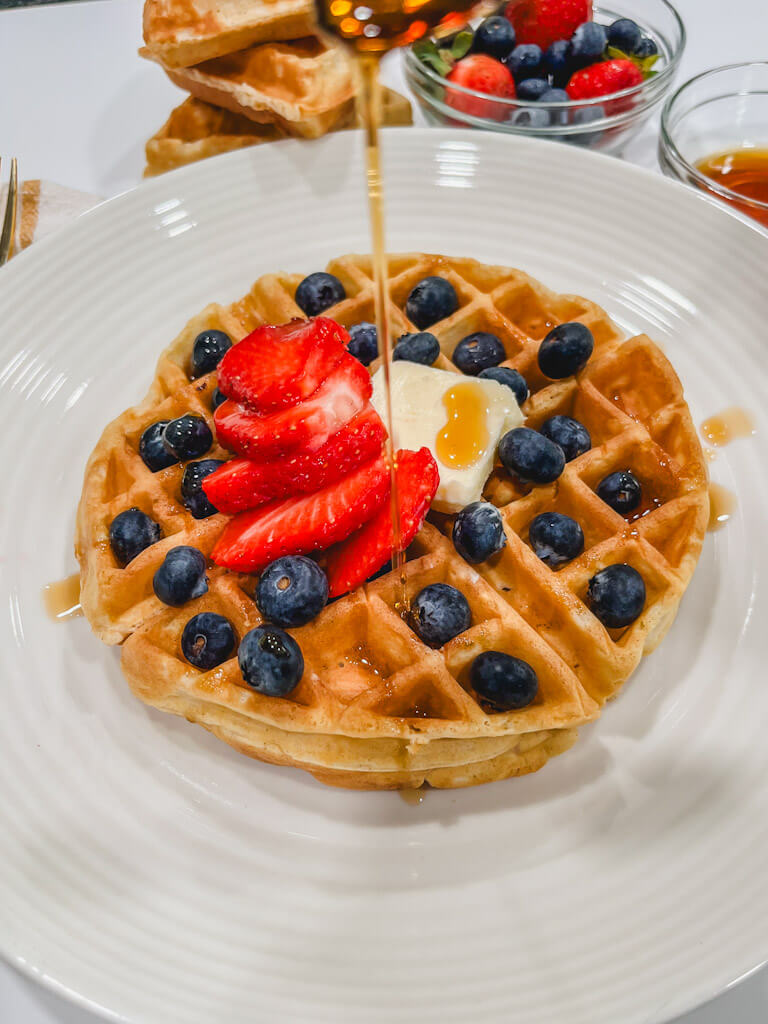 maple syrup being poured onto a waffle with blueberries and strawberries on top.