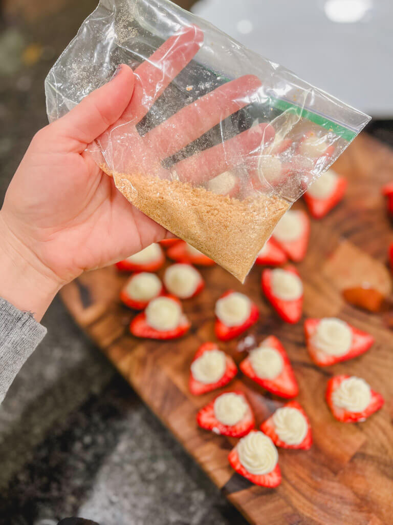 a bag of graham cracker crumbs being held over filled strawberries.