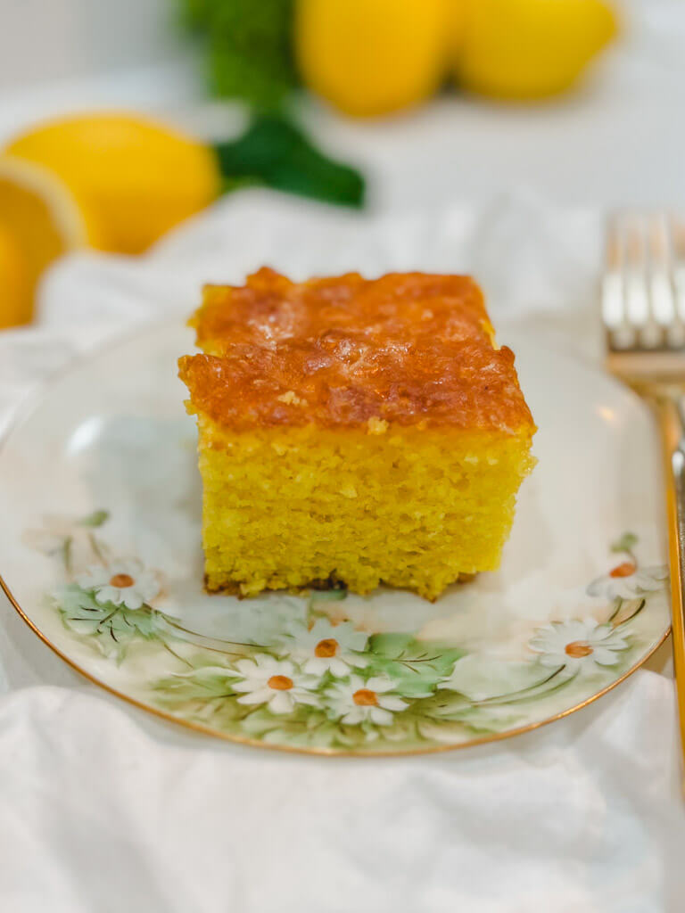 a square slice of lemon cake on a cake with lemons blurred out in the background.