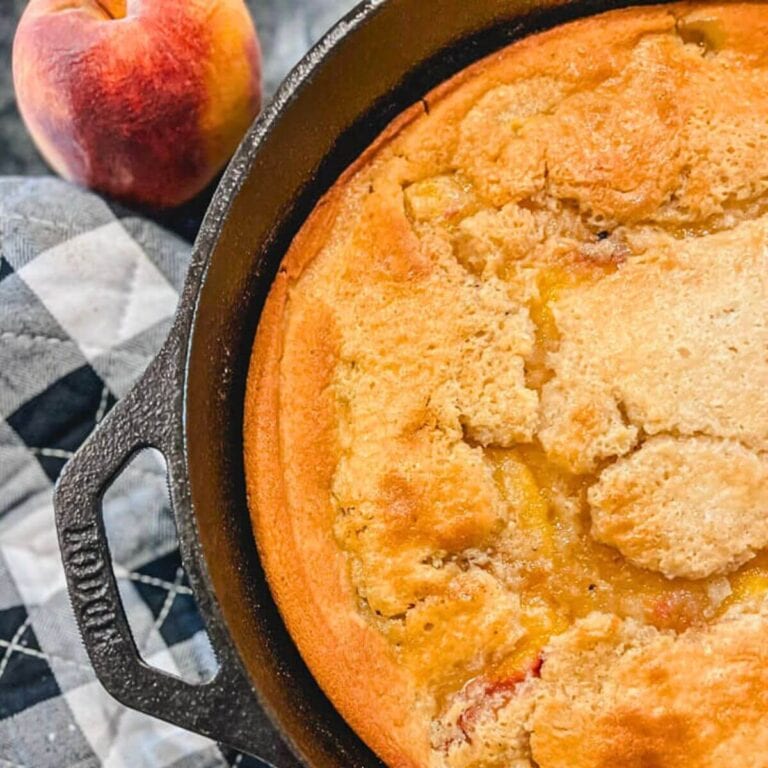 close up of cast iron skillet peach cobbler with a fresh peach next to the skillet.