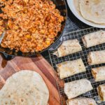 breakfast burritos sitting on a wire rack next to a skillet with the egg mixture and flour tortillas on a white plate and cutting board.