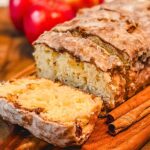old fashioned country apple fritter bread on a wooden cutting board next to cinnamon sticks and whole red apples.