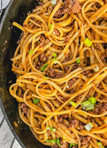 close up of Mongolian beef noodles recipe in a large cast iron skillet next to a black and white kitchen towel.
