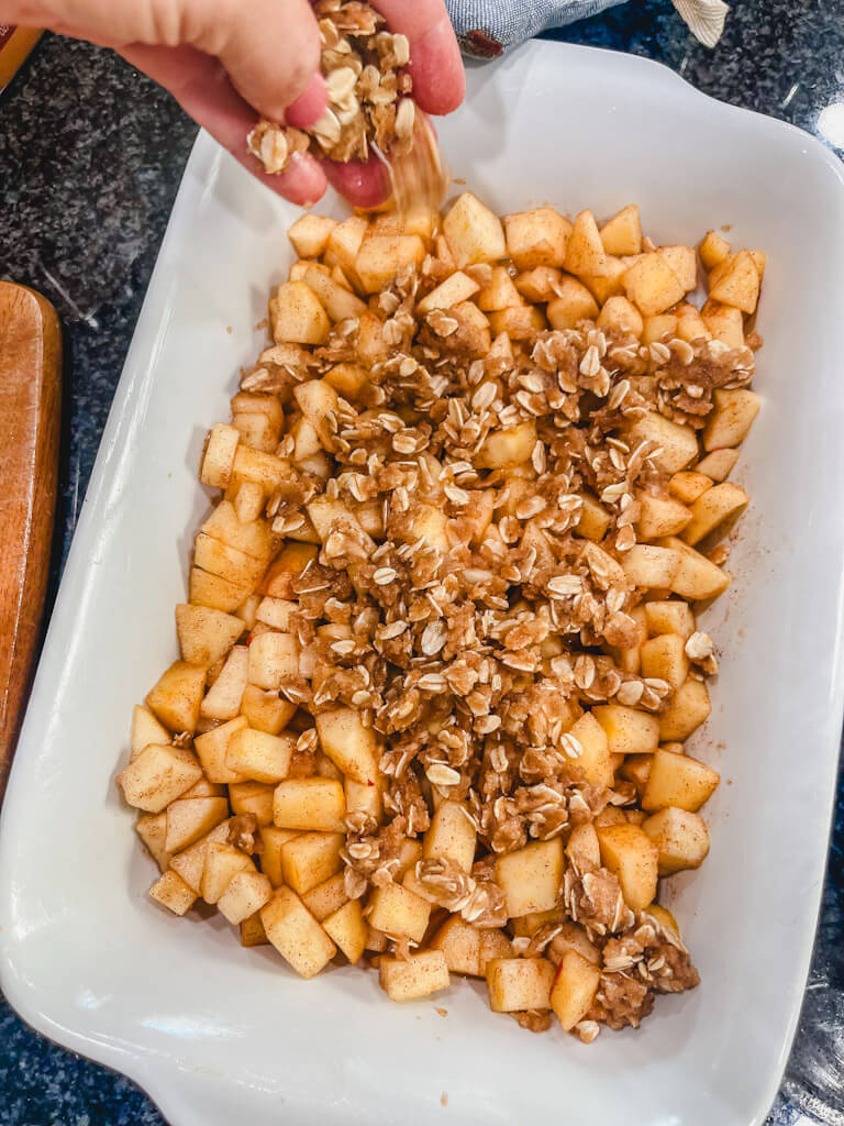 a hand sprinkling the crumble over the tops of the apples in the baking dish for apple crisp recipe.