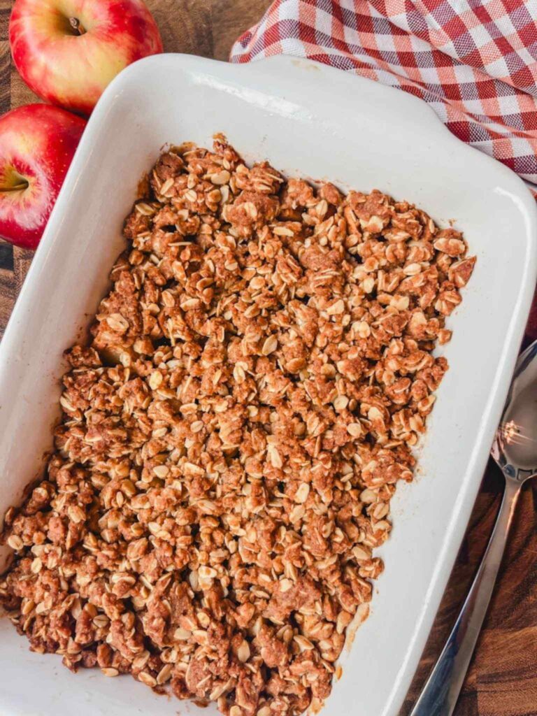 apple crisp recipe with oats in a white casserole dish next to a striped red towel and a couple of whole apples.