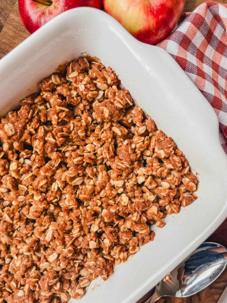 overhead view of apple crisp in a white baking dish next to some apples and a red gingham kitchen towel.