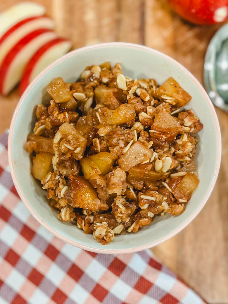 a small bowl holding a serving of apple crisp with sliced apples next to the bowl.
