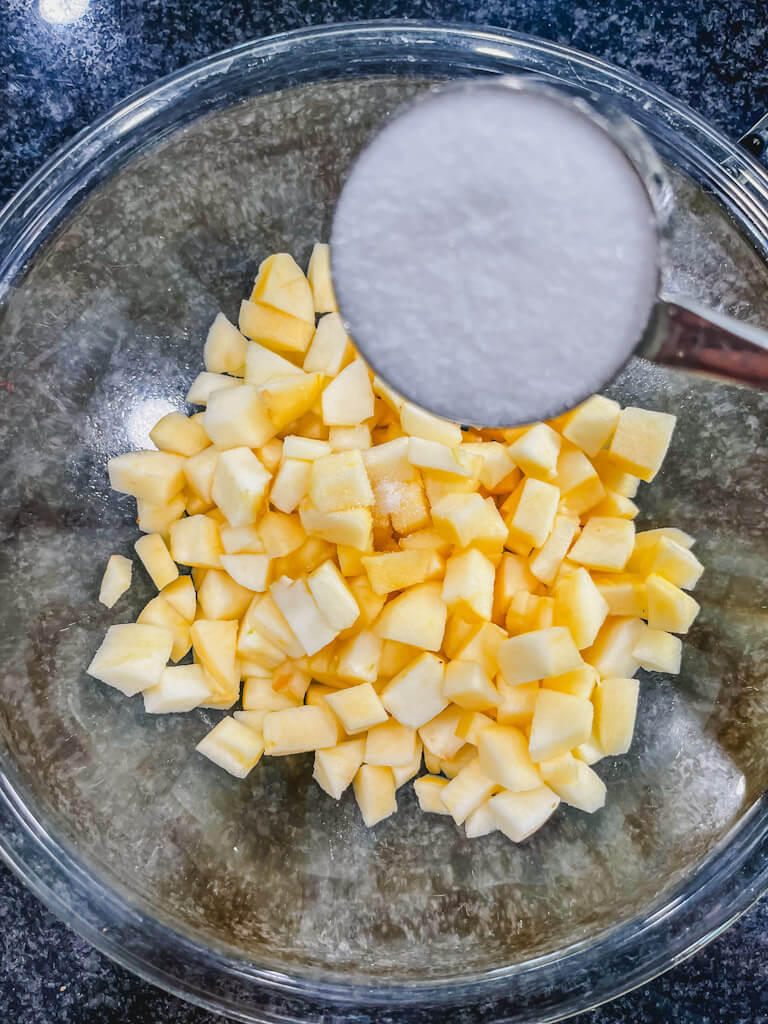 diced apples in a bowl with sugar in a measuring cup being held over the apples.