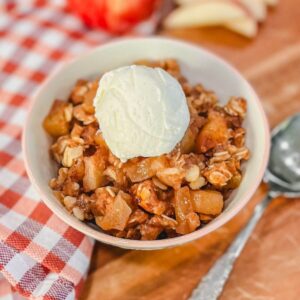 a bowl of apple crisp with oats and a small scoop of ice cream on top next to a spoon and a red striped kitchen towel.