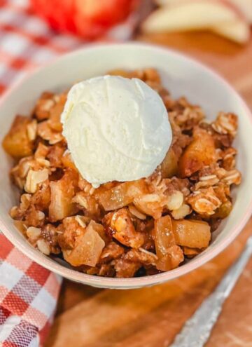 a bowl of apple crisp with oats and a small scoop of ice cream on top next to a spoon and a red striped kitchen towel.