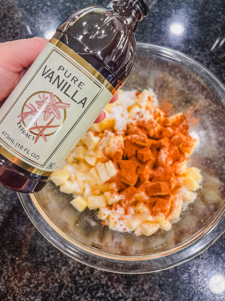 a bottle of vanilla extract being held over the bowl of apples.