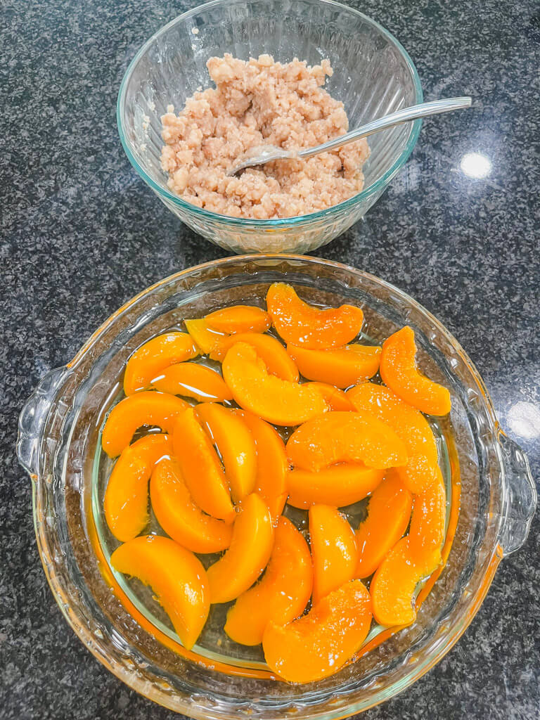 the bowl with the crumble topping and peaches in a baking dish ready for assenmbly.