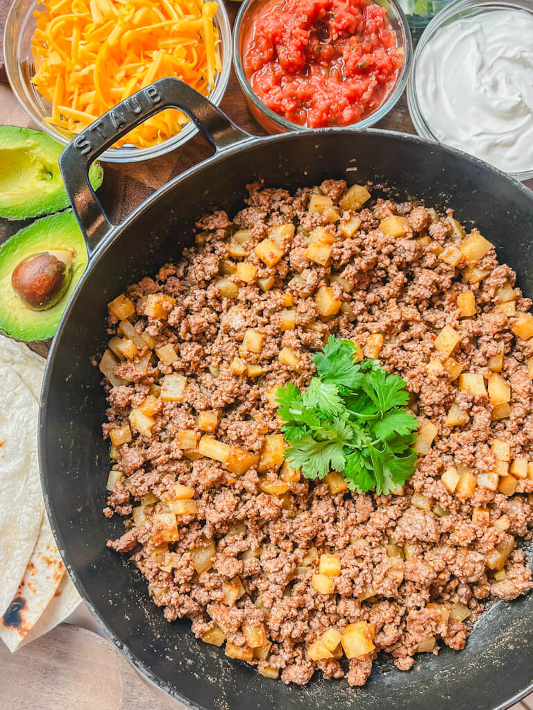 overhead view of beef and potato burrito filling in a skillet next to different toppings.