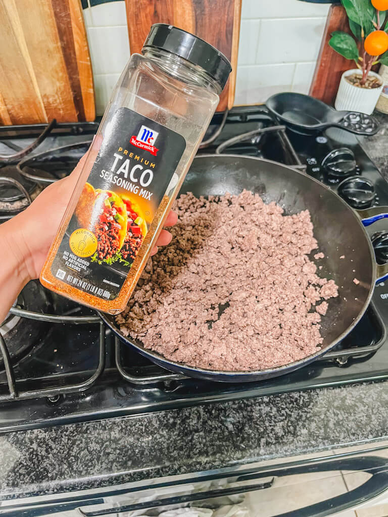a bottle of taco seasoning mix being held over the skillet of ground beef.