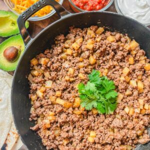 overhead view of beef and potatoes in a skillet next to an avocado, tortillas, salsa, and shredded cheese.