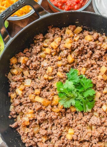 overhead view of beef and potatoes in a skillet next to an avocado, tortillas, salsa, and shredded cheese.