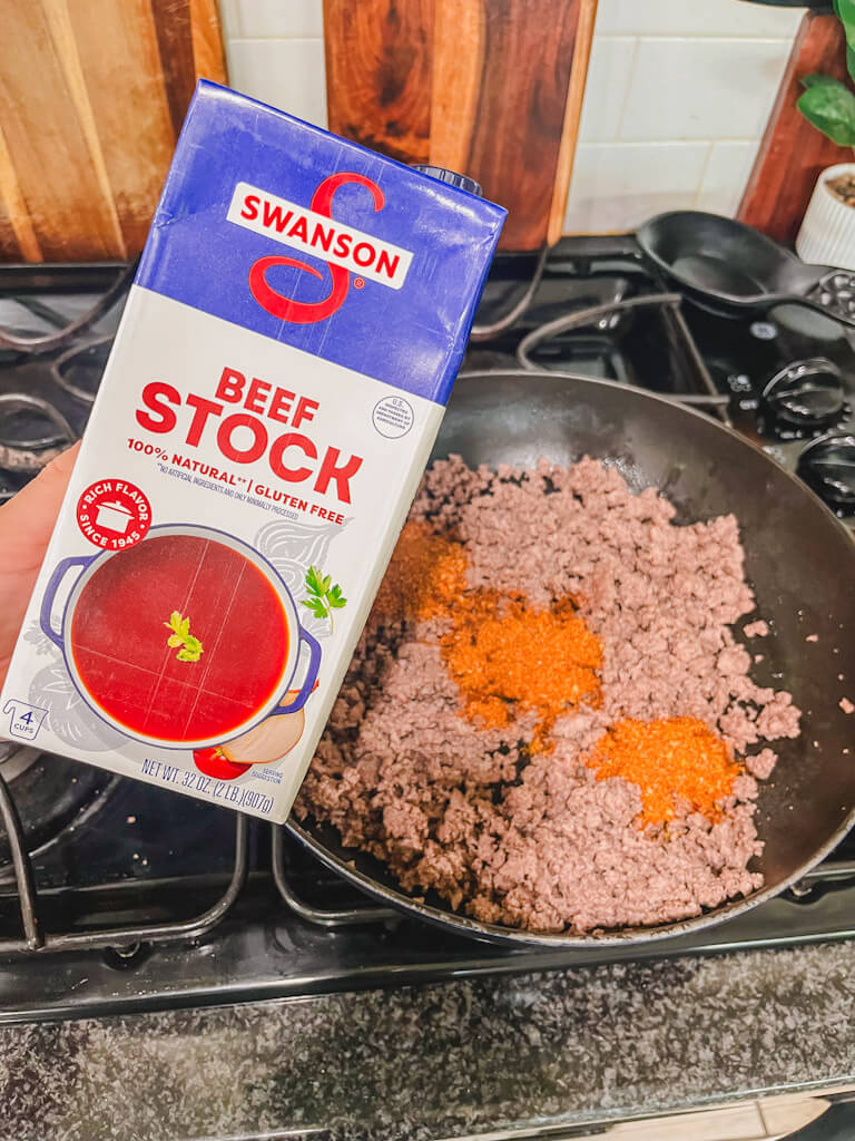 a carton of beef broth being held over the skillet of ground beef.