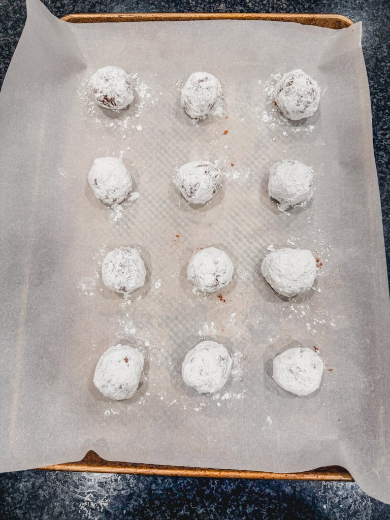 cookie dough balls after being rolled in the powdered sugar once laying on the baking sheet.