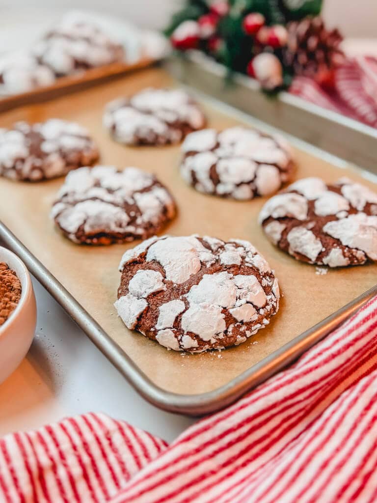 close up of a chocolate crinkle cookie on a baking sheet next to a white bowl of cocoa powder.