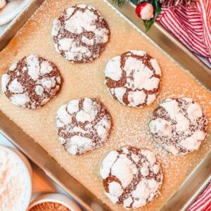 close up of chocolate crinkle cookies on a baking sheet next to a bowl of powdered sugar and a bolw of cocoa powder.