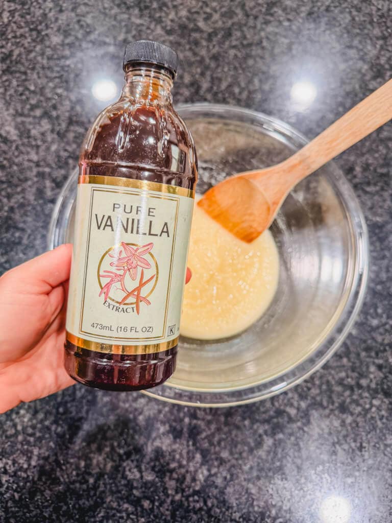 A hand holding a bottle of vanilla extract over the mixing bowl of butter and sugar.