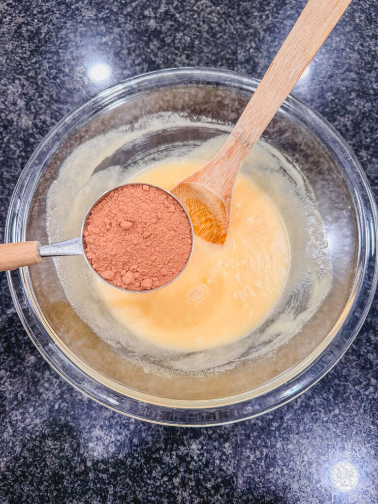 A measuring cup, holding cocoa powder over the mixing bowl for chocolate crinkle cookies.